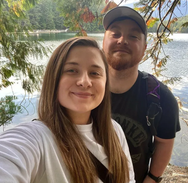 Lexi and Rob smiling in a selfie in front of a forest lake